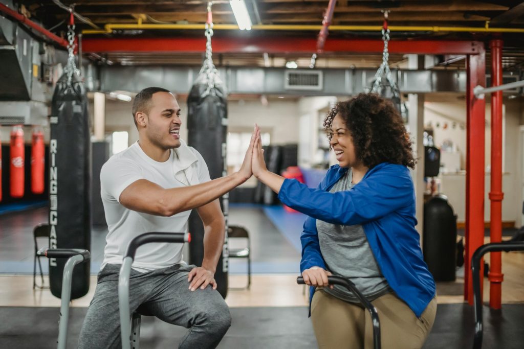 Cheerful plump African American female trainee and fit smiling male coach clapping each others hands while training in spacious equipped gym